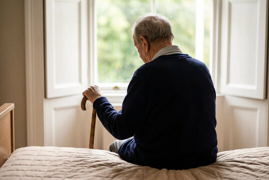 Lonely elderly man sitting on bed holding walking cane near window reflecting aging and isolation
