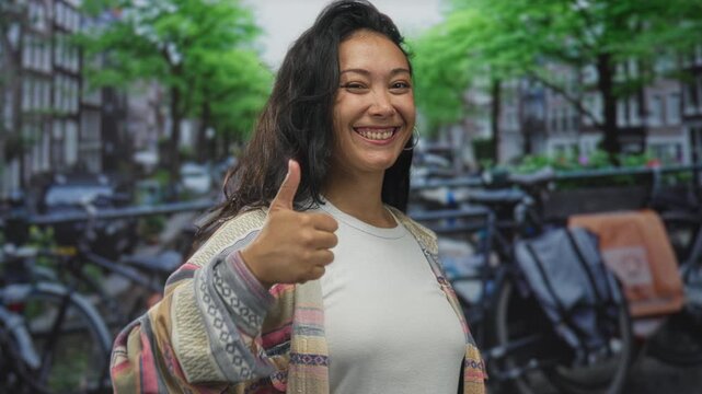 Young hispanic woman giving thumbs up beside parked bicycle rack on city street, smiling broadly; joy confidence.