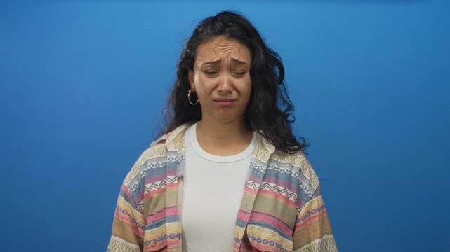 Young hispanic woman grimacing with visible face, raised eyebrow and hoop earrings in a blue studio wearing patterned shirt; disgust.