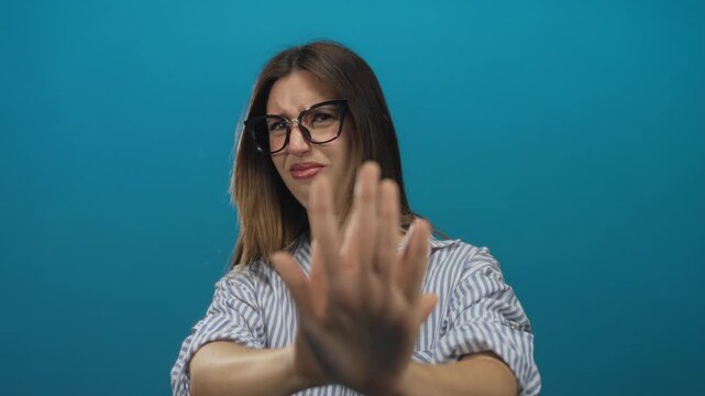 Young hispanic woman in striped shirt wearing glasses pushes palm forward and frowns in blue studio; disgust.