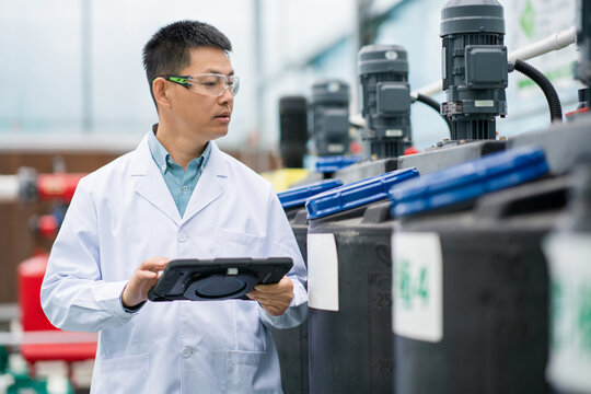 Technical staff in laboratory wearing lab coat and safety goggles checking equipment with tablet computer