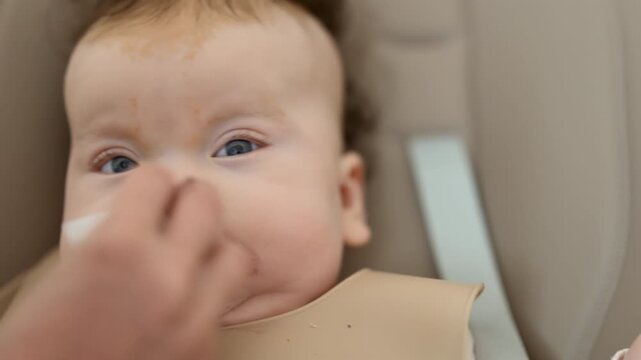 Infant seated in a high chair while an adult feeds the baby with a spoon