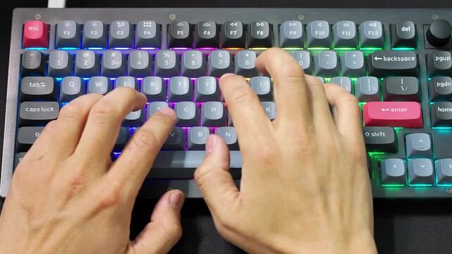 Overhead perspective of colorful gaming keyboard in use. Human hands pressing keys on compact layout device. Concept of esports, creative work, digital communication and modern computing.