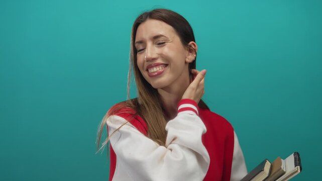 Woman in red and white varsity jacket holds textbooks and tucks hair behind ear in studio; school pride joy.