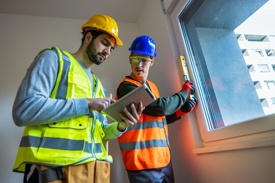 Two construction workers using a digital tablet to check window installation and alignment in a new apartment.