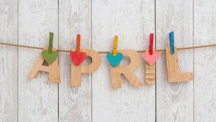 Wooden April letters with colorful hearts and clothespins on a string against white wooden background