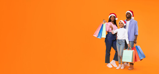 A family stands together against an orange background, wearing Santa hats and holding shopping bags. They appear happy and excited about the holiday season. © Prostock-studio
