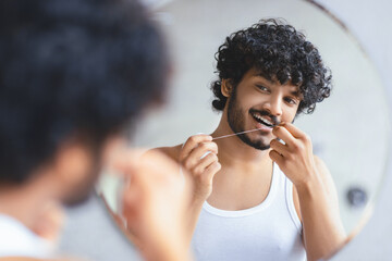 A young man is seen looking in the bathroom mirror while flossing his teeth. He stands in front of a round mirror and wears a white tank top. The setting is bright with light tones. © Prostock-studio