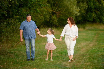 Fototapeta premium Family walks together in park in fresh air, strengthening bonds during holidays and vacations. Mom, dad, daughter walk through park holding hands and chatting joyfully. Parent and child, memories.