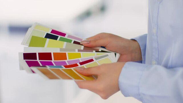 A person with light skin holds a set of colorful paint swatches in a design studio. The workspace is bright and showcases various colors for a creative project.