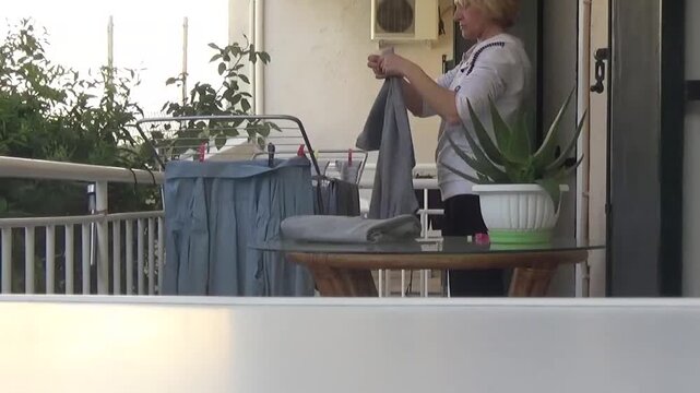 A young woman collects laundry from a drying rack on the balcony. A woman folds dry sweatpants from the drying rack. A beautiful potted cactus sits on a glass table, reflecting the rays of the spring 