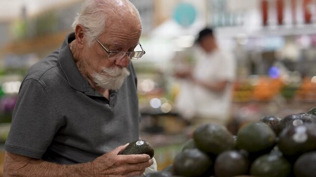 Senior man is shopping for food in grocery store and checking ripe avocados while preparing to bag his selection.