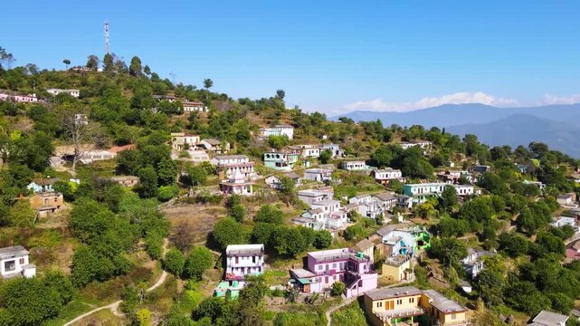 Aerial drone view of a scenic rural village. Eagle eye shot of nestled green hills, Garhwal Uttarakhand India. Houses terraced slopes winding mountain roads and clear blue sky. Natural Himalayan lands