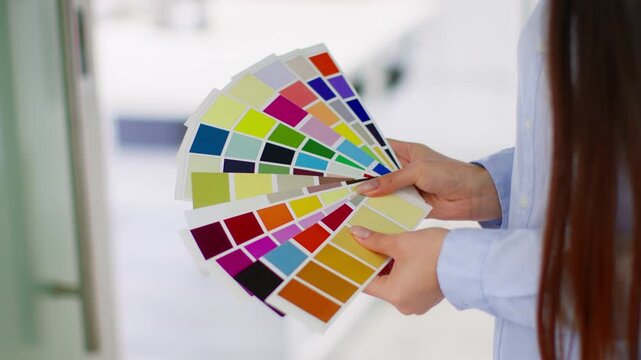 A woman with long dark hair is holding multiple color swatches in a light and bright room. She appears focused on making choices for a design or decorating project.