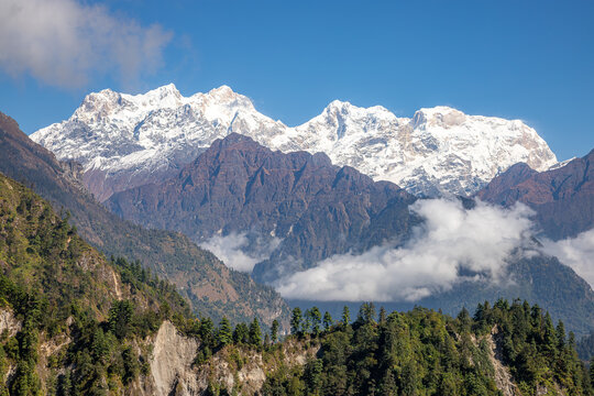 Snowy Himalayan massif Manaslu rises above dark ridges and drifting clouds under a clear blue sky, Annapurna Circuit trekking route, Nepal.