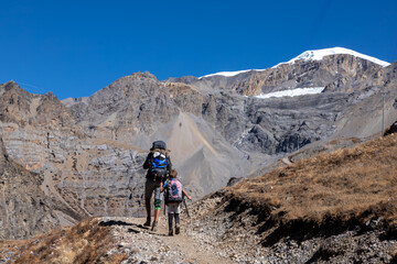 Two hikers, an adult and child, walk a rocky high-altitude trail toward barren Himalayan cliffs and snowy ridges, Annapurna Circuit, Nepal. © Max Maximov