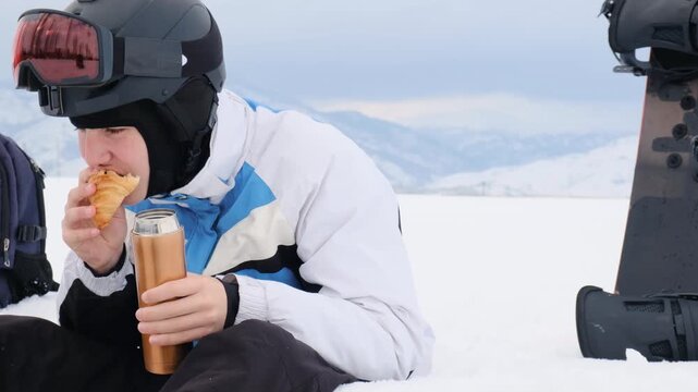 Boy snowboarder bites croissant with chocolate filling and drinks hot tea from thermos while sitting on snow after active snowboarding. Increased appetite in fresh mountain air after physical activity