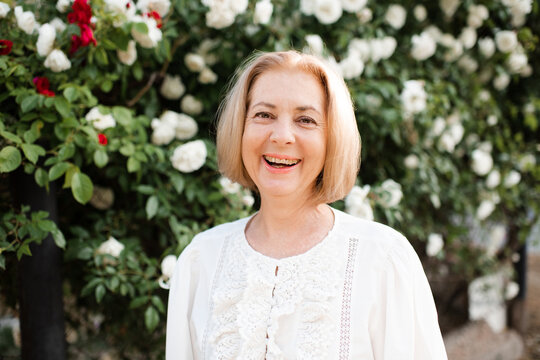 Smiling senior woman 60-65 year old with short blonde hair posing over blooming rose bushes outdoors. Wellness lifestyle.