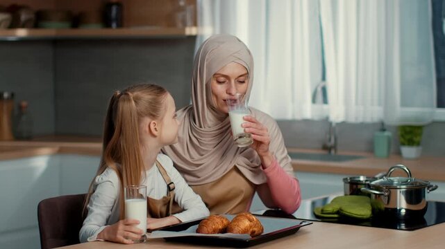 A woman in a hijab sits at a kitchen table with her daughter. They smile at each other while sharing croissants and glasses of milk. The kitchen has modern decor and natural lighting.