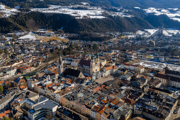 Obraz premium Aerial view of Brixen, Italy, shows Brixen Cathedral towers, St. Michael spire, cloister, pastel townhouses, terracotta roofs, crisp light, fields, and forested hills.