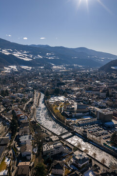 A bright aerial view of Brixen, Italy, shows the Eisack River, twin towers of Brixen Cathedral, and a circular modern complex with vents under a clear winter sky.