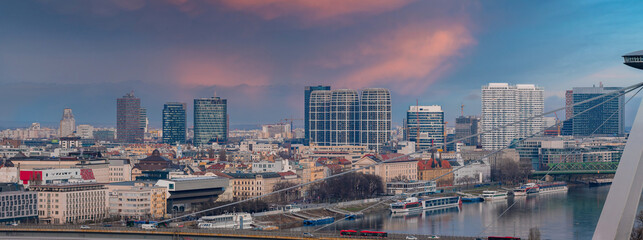 Obraz premium Aerial view of Bratislava, Slovakia, at dusk, with the Danube River curving, riverboats moored, modern towers and historic blocks, and the UFO Bridge cables at right.