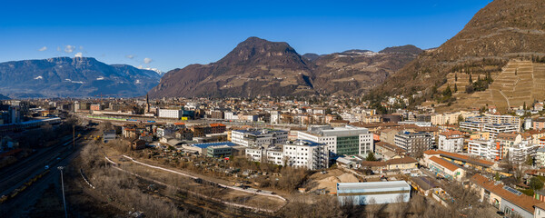 Fototapeta premium A wide aerial view of Bolzano, Italy shows Monte Tondo, terraced vineyards, a rail yard, pastel blocks, offices, and a church spire under crisp winter light and clear skies.