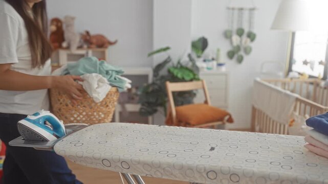 Young woman holding a laundry basket in nursery near ironing board and crib indoors; tired patience.