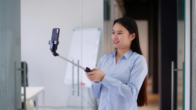 A young Asian woman stands in a brightly lit office, smiling while using a selfie stick to take a photo of herself. She enjoys her time while engaging with her smartphone.