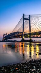 Obraz premium Suspension bridge over water, illuminated against a dusk sky, with stone embankment foreground