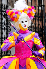 Obraz premium Venice, Italy - February 09, 2026: Colorful masked performer in vibrant costume with floral headdress poses during Venice Carnival celebration in front of ornate wrought iron gate