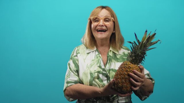 Woman holding ripe pineapple with both hands, smiling and laughing with teeth visible while wearing leaf print shirt in studio; joy tropical.