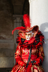 Obraz premium Venice, Italy - February 09, 2026: Elegant woman in ornate red costume and mask with feathers poses against a column during Venice Carnival celebration in Italy