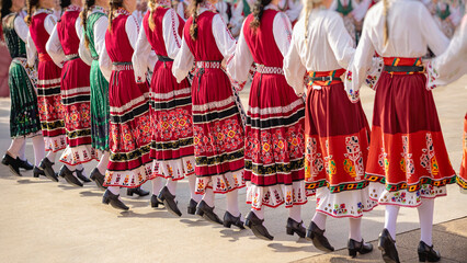 Naklejka premium Women in traditional embroidered folk dresses dancing in synchronized movement at cultural festival. Bulgaria heritage, national identity, folk horo dance tradition, artistic craftsmanship of region