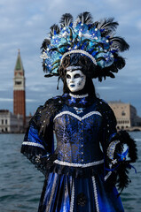 Obraz premium Venice, Italy - February 09, 2026: Woman in elaborate blue costume and mask with feathers poses by water during Venice Carnival, iconic bell tower visible in background