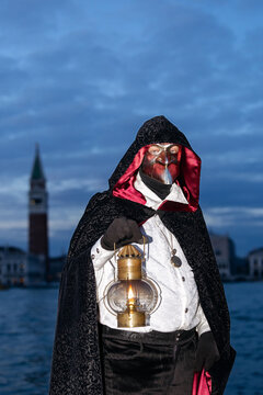 Venice, Italy - February 09, 2026: Man in black cloak and mask holding lantern during Venice Carnival with historic bell tower visible in the background