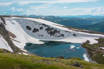 Scenic sunlit landscape with alpine lake in rocky snowy cirque near stone hill top during thaw. Ice floats in mountain lake among rocks and snows with view to forest mountain range under cloudy sky. © Daniil