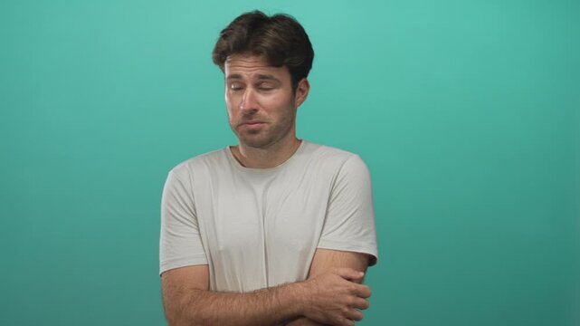 Man holding his upper arm and looking to the side in a teal studio setting with bare arms and a neutral tshirt; uncertainty hesitation introspection.