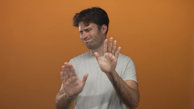 Man in white tshirt holding both palms out stop gesture in orange studio; rejection boundaries caution.