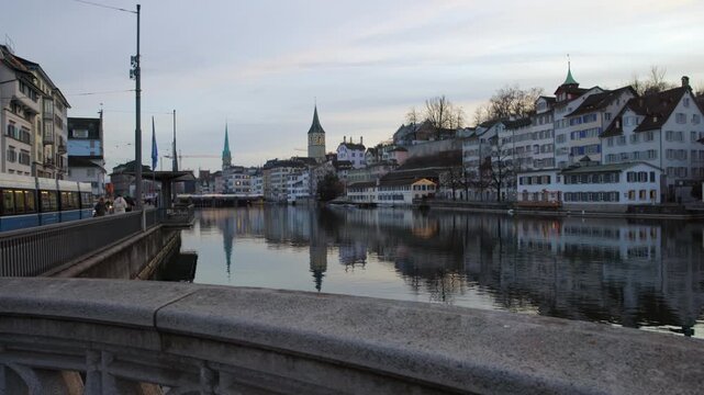 Panning shot of the Limmat river in Zurich, Switzerland, showcasing the historic old town buildings, Fraum&uuml;nster church, and St. Peter's tower reflected in the calm water at dawn.