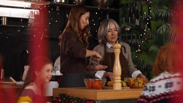 A grandmother chews and listens to her daughter talking while they stand at a table with her grandson and granddaughter running around