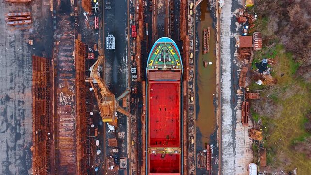 Overhead view of a ship hull in construction. Top down shot of a red and blue ship hull positioned between industrial cranes and metal structures.