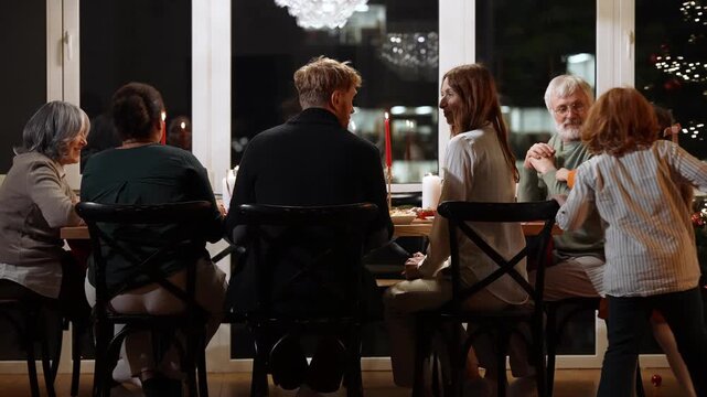 Family and guests sit at a table and talk while a grandson and granddaughter run and hug their grandfather