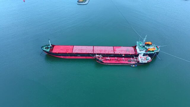 Cargo ship and small tanker vessel side by side on water. Aerial view of a large red and black cargo vessel moored next to a smaller bunkering tanker on open water.