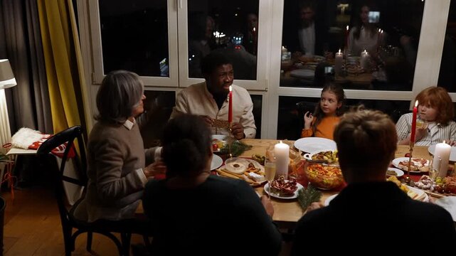 A male guest and a grandmother sit at a table with family and guests while they talk and eat