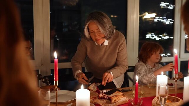 A grandmother stands while cutting a pie and placing a piece on a plate on the table held by a male guest sitting next to her