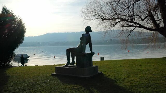 Panning shot of the "Sitzende Badende" bronze sculpture at Zuerichhorn park, situated on the green lakeside of Lake Zurich with mountains in the background.
