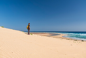 Woman hiking on sand dunes overlooking a vast beach in Fuerteventura, Canary Islands, Spain, enjoying panoramic ocean views under a clear blue sky.