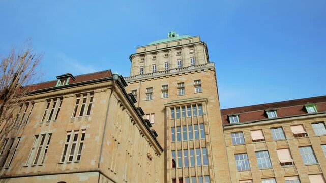 Low-angle panning shot of the University of Zurich (UZH) main building, featuring its historic stone facade and the prominent tower with a green dome under a clear blue sky.