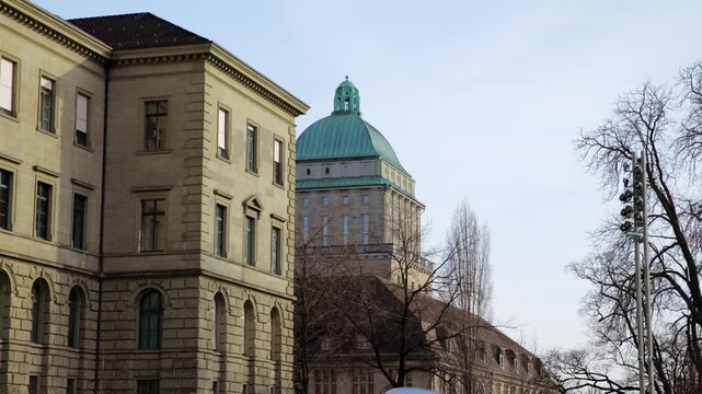 Panning shot of the University of Zurich main building, starting from the neoclassical stone facade and moving to the iconic green copper dome against a clear blue sky in Switzerland.